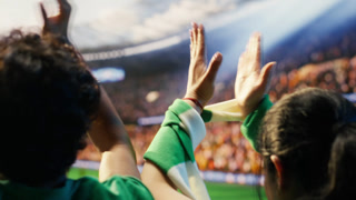 Close up of football supporters clapping across packed stadium seats and beating drum. Rear view of soccer fans applauding during loud match