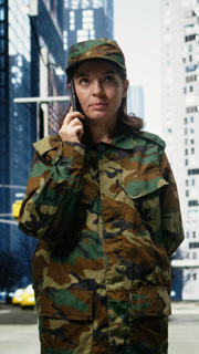 Vertical video Female soldier making call on urban street outdoors in metropolitan downtown. Woman wearing camouflage uniform speaks on