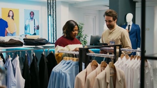 Store assistant helping client with clothes, shopping for trendy collection. Young adult talking to mall employee about fashionable merchandise, fashion boutique.