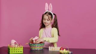 Cute smiling toddler decorating a basket with painted easter eggs, presenting her handcrafted festive arrangement in studio. Happy lovely