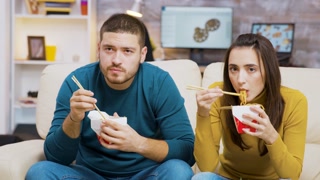 Bearded man and his girlfriend eating noodles with chopsticks while watching tv.