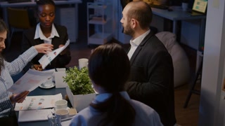Diverse multi ethnic businesspeople sharing management presentation on paperwork discussing company strategy. Teamwork brainstorming ideas while working in meeting room late at night