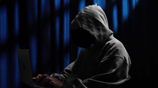 Anonymous hooded hacker programming on laptop computer