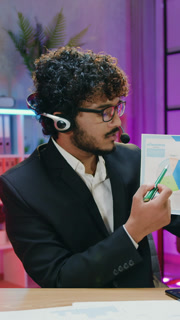 Business concept where likable man office manager in wireless headphones talking into camera in night lighted workroom and presenting