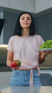 Beautiful smiling 30-aged brunette standing in cozy cuisine and doing choice between sweet mango and green fresh lettuce,front view