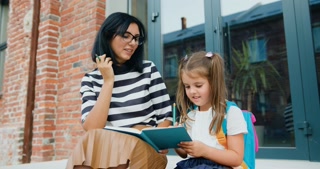 smiling young mother and her happy little daughter with backpack using pencils writing something in a notebook while sitting on