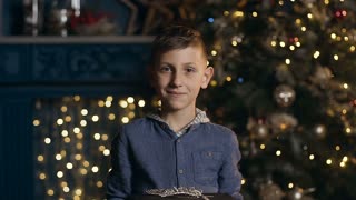 Brown hair boy smiling and giving christmas gift to the camera on the christmas tree and boken from light. Close up shot.