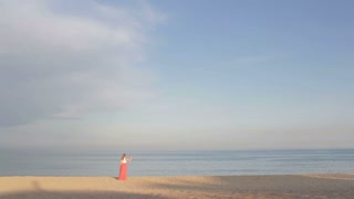 Girl in orange dress says good bye to the sea and is walking away. Wide shot. Shot in golden hour. Nice warm sunset colors.