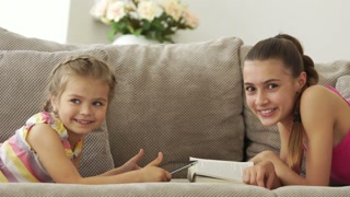 Two sisters lying on the couch with a book and a tablet pc