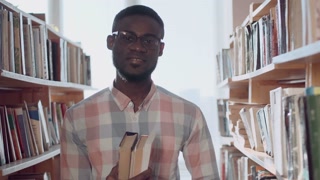 Young man student in glasses exploring library bookshelves, smiling and showcasing closed books