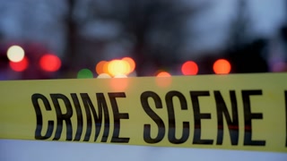 A bright yellow Crime Scene Do Not Cross tape stretched across an urban environment, marking a restricted area for investigation. Flashing