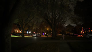 Nighttime suburban neighborhood street with Halloween decorations and glowing lights in residential yards. Cars driving through a quiet