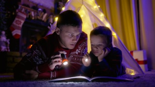 Dad with son in canvas tent at home. Young handsome man with flashlight highlighting the book and reading the fairytale story to small boy