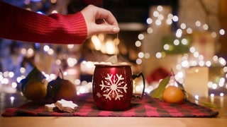 Female boiling hot tea in holiday themed mug. Hand with tea packet in red glossy mug with snowflake on wooden table with plaid napkin