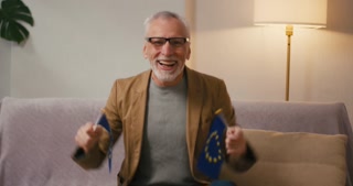 Mature Arab man says Yes waving European Union flags sitting on sofa. Bearded retired man in jacket expresses satisfaction with political