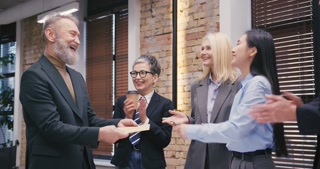 Young Asian woman provides certificate to senior bearded man. Multiethnic businesspeople shake hands while their colleague stand in the middle smiling.