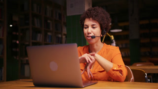 Beautiful African American woman wearing headphones with microphone using laptop, having video call, sitting in modern coworking. Online