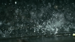 Splashing raindrops ripple across dark reflective water surface in slow motion