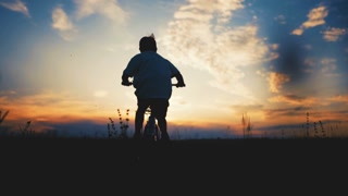 Silhouette of carefree boy riding bike in lawn, moving to horizon in sunset. Happy child riding and enjoying life, freedom and summer