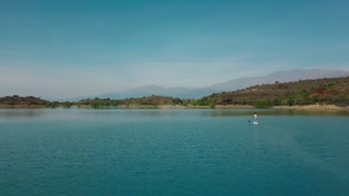 Aerial drone flying over a man doing stand up paddle in a blue water lake with mountains landscape