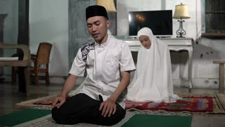 Asian father and daughter praying indoors, quiet living room prayer scene with man in cap and young girl in white veil on carpet