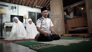 Asian young imam guiding sisters in prayer, male instructor seated on mat with two girls behind in white prayer garments, rustic