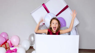 Cute, joyful little girl in pink dress jump out of the big present gift box with balloons at home birthday party streamers, Happy birthday. Celebrating