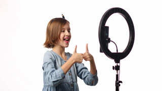 Smiling little girl blogger in casual denim dress use selfie led lamp and smartphone making online video, vlogger live stream isolated on white background, studio. Excited face, lifestyle concept