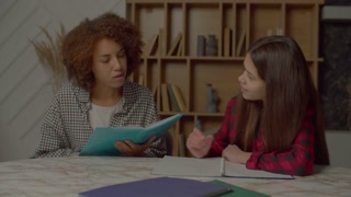 Caring charming African American mother with tutorial reading school assignment, helping smart multicultural teenager daughter to do homework while family studying together in domestic room.