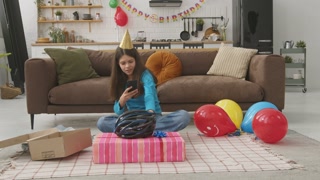 Joyful cute teenage girl in party hat sitting on floor, taking photo of present on smartphone and sharing post in social network about birthday gift, expressing excitement and happiness at home.