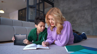 Portrait of caring pretty teenage nanny and cute school age black boy studying together , solving mathematical example and writing solution into exercise book while lying on floor at home.