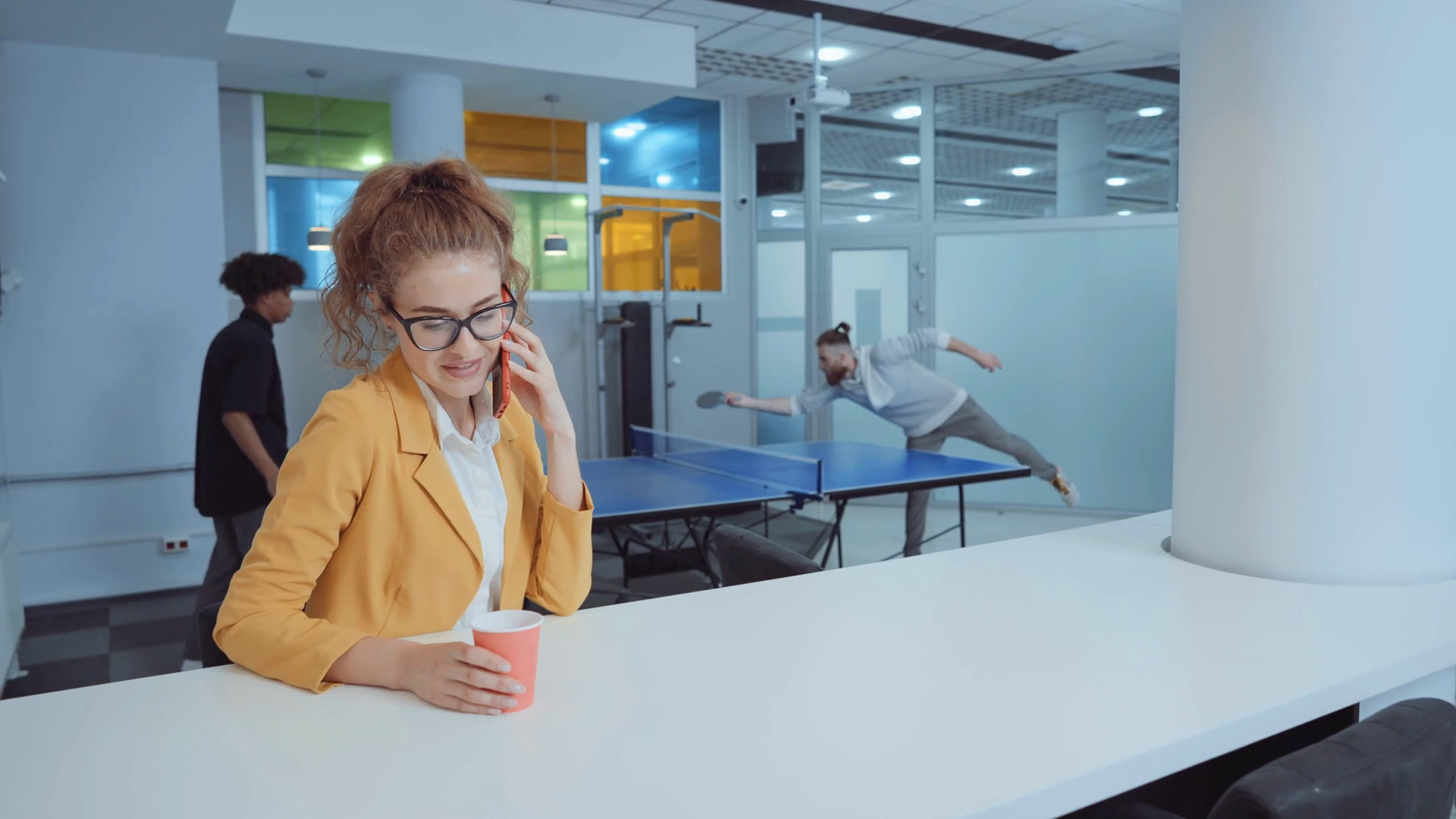 Cheerful woman in eyeglasses sitting in lounge zone and talking on phone, coffee cup in her hand