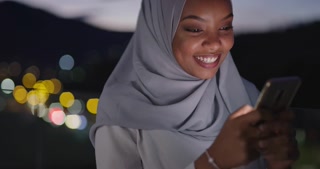 Muslim woman in scarf texting on smartphone at night in city with bokeh lights