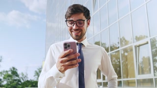 Young professional smiles while using smartphone outside a modern glass office building on a sunny day