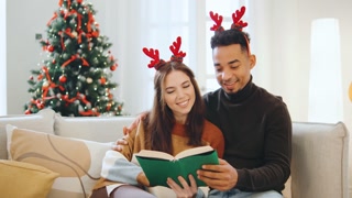 Multiracial couple enjoying a cozy evening by the Christmas tree while reading together this festive New Year
