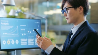 In the Office Asian Businessman Uses Smartphone, Typing Important Message. His Desktop Computer Shows Sratistics with Company Growth. Stylish Office.