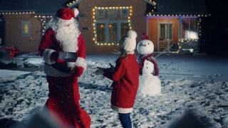 Magical New Year Evening, Authentic Santa Claus Gives Cute Little Girl a Wrapped Gift Box. In the Background Idyllic Decorated Front Yard, with Snowman, Christmas Tree and Idyllic House.