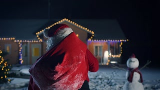 Authentic Santa Claus Tosses Red Bag Over His Shoulder and Walks into Front Yard of the Idyllic House Decorated with Lights and Garlands. Magical New Year's Eve with Falling Snow.
