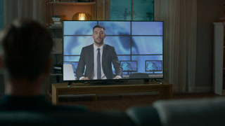 Man Watches Evening News on TV while Sitting on a Couch at Home in the Evening. Cozy Living Room with Warm Lights. Over the Shoulder Shot.