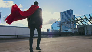 Businessman Superhero With Red Cape Blowing in the Wind Walks on the Roof of a Skyscraper Ready to Save the Day. In the Background Modern City Center. Following Back View Shot.