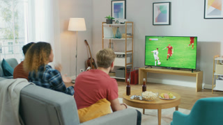 Three Sports Fans Sitting on a Couch in the Living Room Watch Important Soccer Match, Worry for For their Team. Bright Cozy Apartment with Friends Eating Snacks and Having Fun.