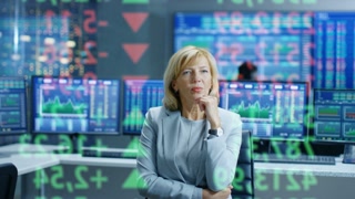 Stock Market Female Top Manager Looks at Projected Ticker Numbers and Graphs. Behind Her Room Full of Screens and Statistics.