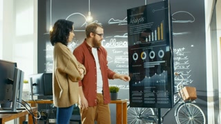 Female Developer and Male Statistician Use Interactive Whiteboard Presentation Screen to Look at Charts, Graphs and Growth Statistics. They Work in the Stylish Creative Office.