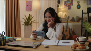 Medium shot of gen Z Asian girl watching video on smartphone online while doing break during homeschooling