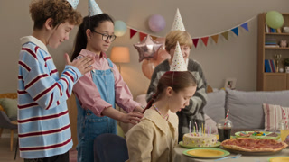 Three friends with party hats standing behind birthday girl who sitting at table and blowing out candles on cake