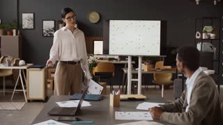 Brunette mature woman standing near whiteboard, explaining statistics while her biracial colleagues sitting at table with laptop and reports