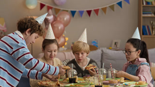 Group of diverse friends in party hats gathering around table and enjoying pepperoni pizza with gooey cheese during birthday celebration