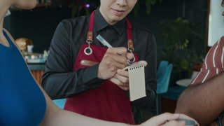 Tilt down shot of Asian waiter making notes while listening to guests ordering dinner at restaurant