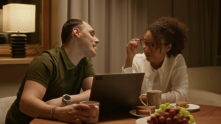 Flirty biracial couple kissing each other while eating grape. They sitting at table, using laptop and enjoying romantic atmosphere