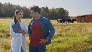 Young female veterinarian in white coat shaking hands with male farmer after consultation on livestock health, standing in green field with cows grazing nearby on organic dairy farm, slow motion